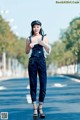 A woman in overalls and a hat is standing on the street.